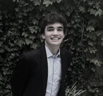 A person in a suit, smiling in front of leafy background.