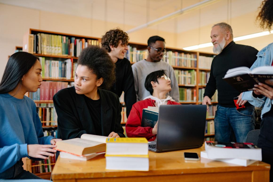A diverse group of six individuals collaborates in a library, with two young women seated at a wooden table and four others standing nearby, engaging in conversation. A laptop and several books are on the table.