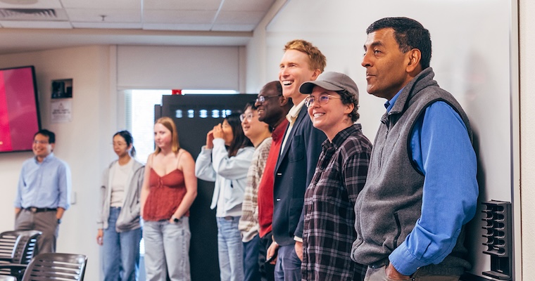 A group of faculty and students standing up against a white board on the right side of the frame, looking off frame to the left with smiling expressions.