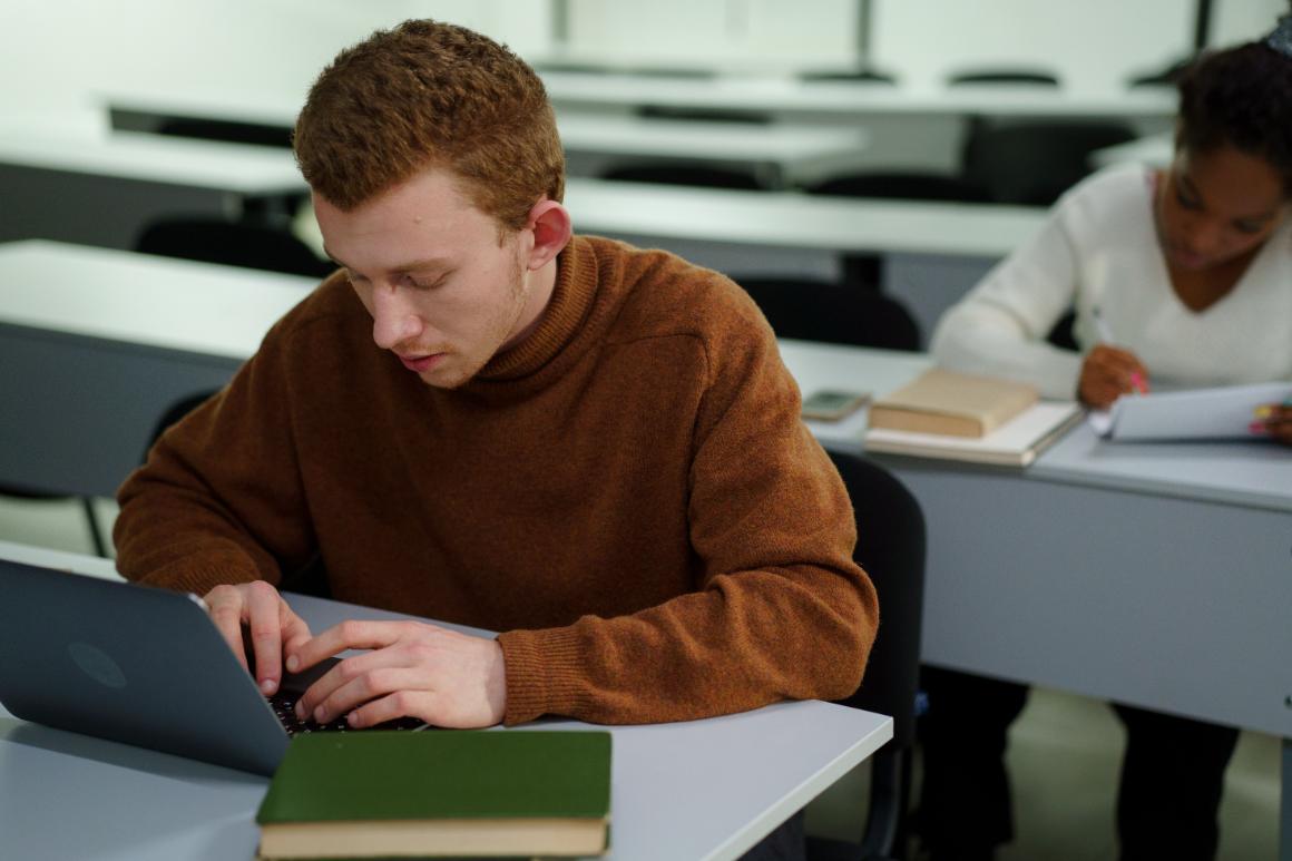 Two students studying in a classroom; one is typing on a laptop while the other writes in a notebook.