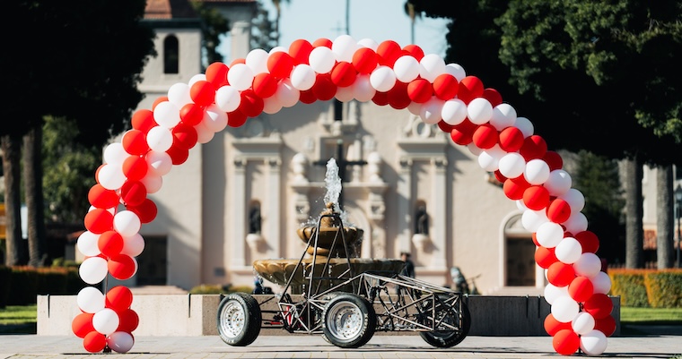 Bronco racing car beneath a red-and-white balloon arch in front of a mission-style building.
