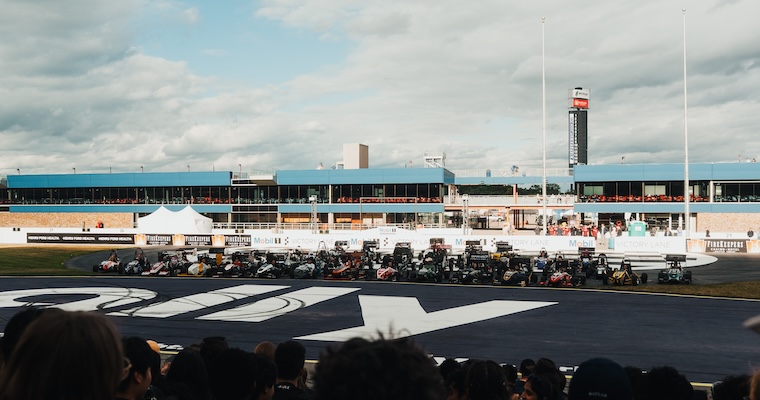 Formula SAE race cars line up across a track as spectators watch from the foreground.