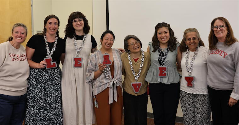 A diverse group of eight L.E.A.V.E.Y. award winners poses together, holding letters that spell L.E.A.V.E.Y. while wearing large chain necklaces.