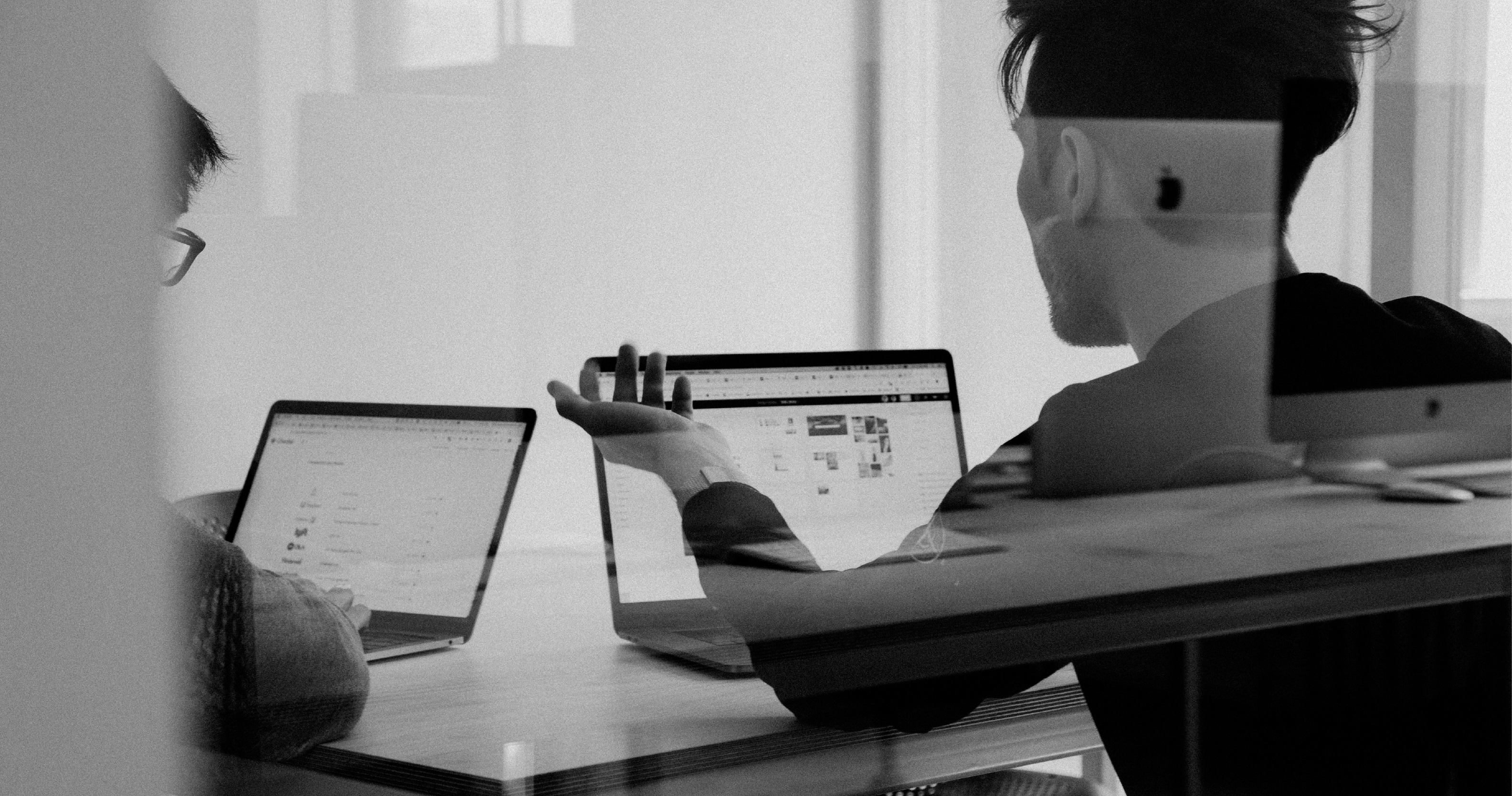 Two people are in an office setting, focused on laptops. One is gesturing towards a screen displaying a website layout.