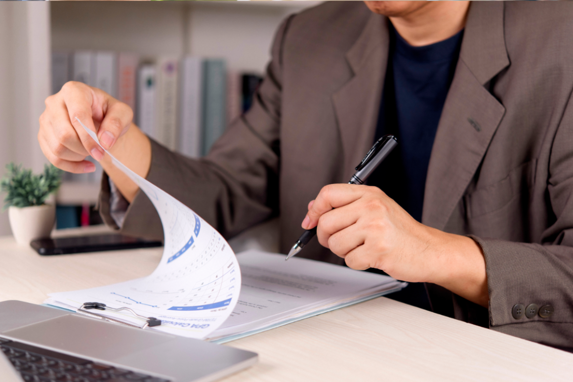 A person in a suit holding a sheet of paper while writing with a pen on a desk with a laptop and a potted plant in the background.