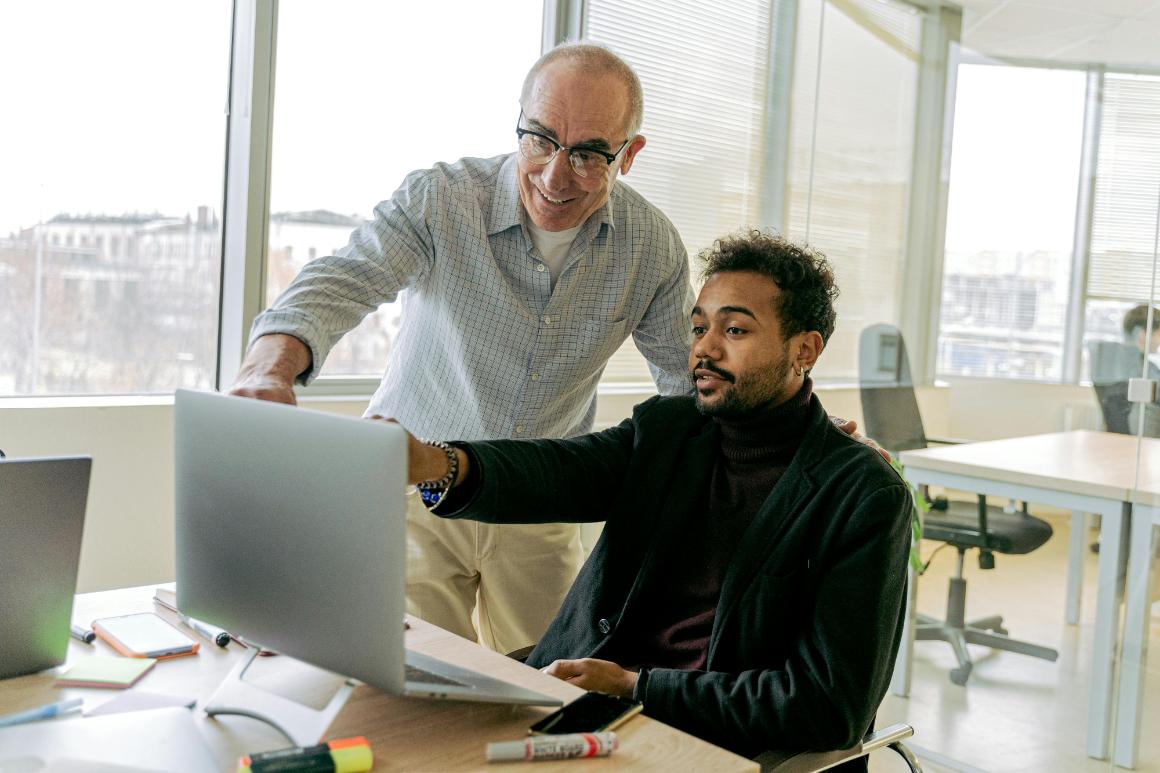 An older man stands and points at a laptop screen while a younger man sits and looks at the screen in a bright office setting.