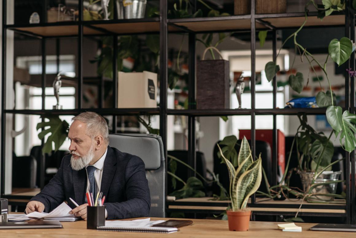Bearded man in a suit reviewing papers at a desk in a plant-filled office.