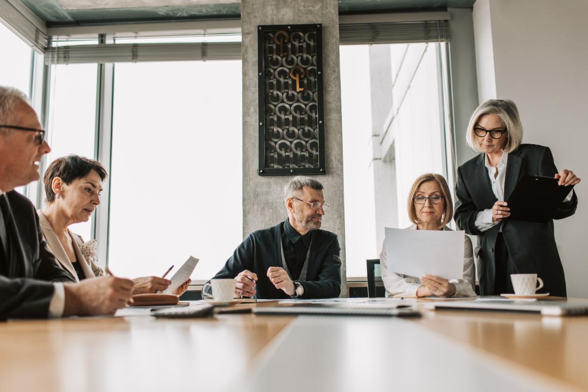 Five business professionals are seated at a conference table reviewing documents, with one standing and holding a clipboard in a modern office setting.