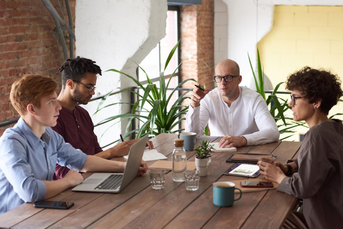 Four adults meet around a wooden table in a bright loft, with one man gesturing while others listen.