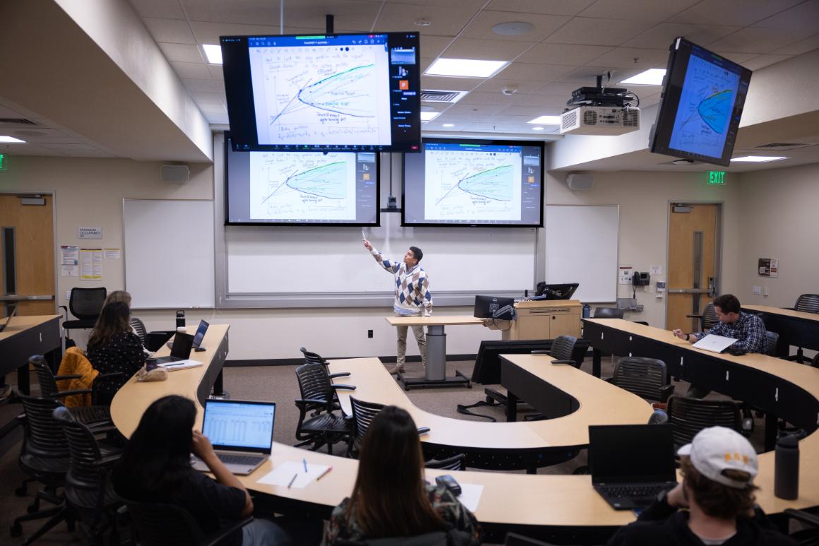 A lecturer in a sweater points at a graph on a large screen in a modern classroom. Several students sit at curved desks with laptops, attentively listening.