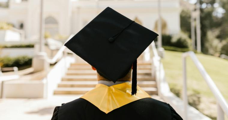 A graduate in a cap and gown with a yellow stole stands facing a sunlit staircase, symbolizing new beginnings and achievement image link to article