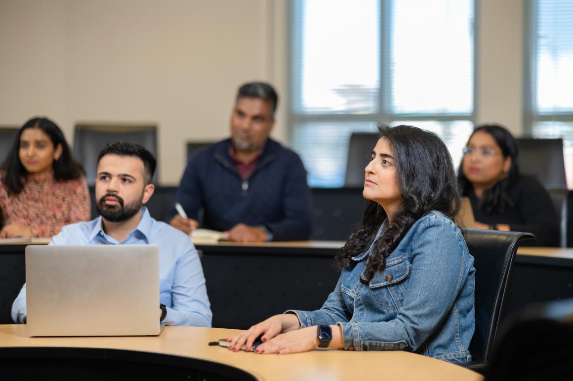 A group of MBA students attentively listening in a bright classroom, with one man at a laptop and a woman in a denim jacket looking upwards.