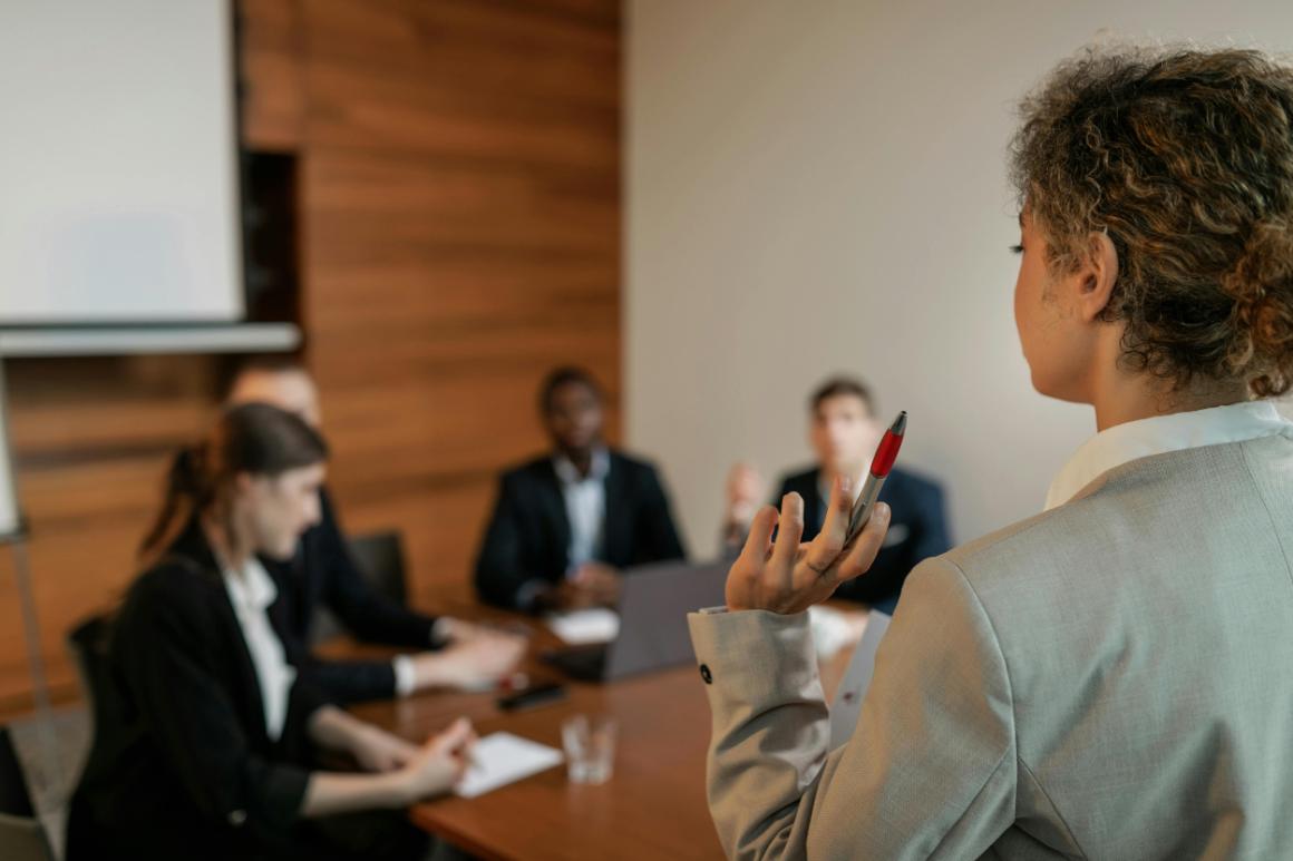 A woman in a gray suit gestures with a pen during a business meeting, while four individuals are seated around a wooden table in a modern conference room.