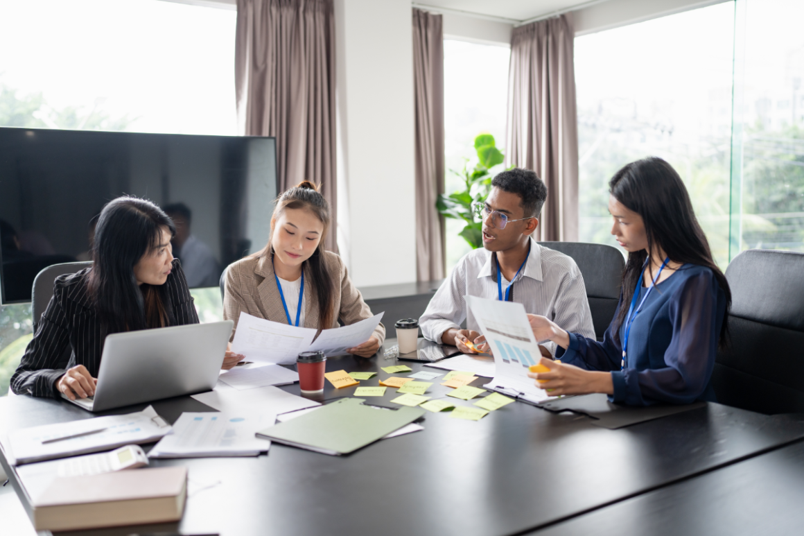 Four coworkers review documents around a conference table in a bright office.