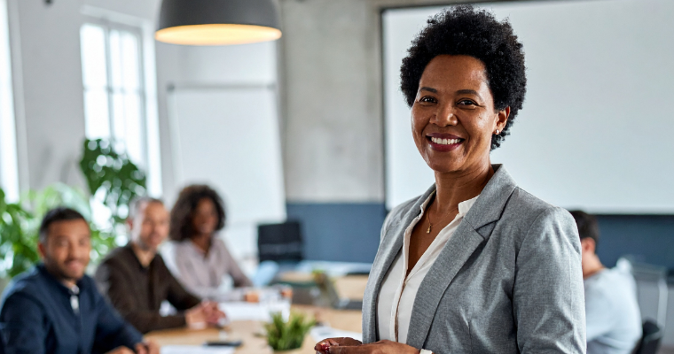Smiling woman in a gray blazer standing in a modern conference room with colleagues seated at a table behind her.