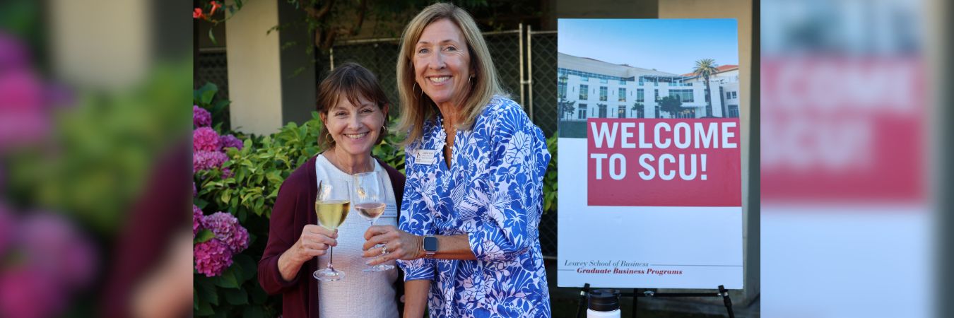 Two admissions advisors toast with wine glasses outdoors beside a Welcome to SCU! sign.