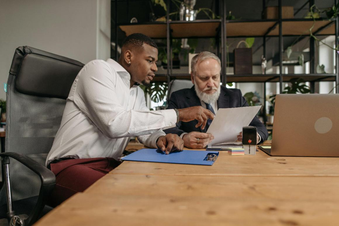 Two men in a modern office discuss a document at a wooden table, surrounded by greenery and office supplies.