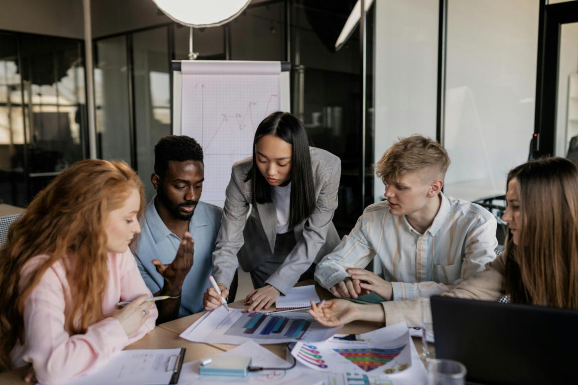 A diverse group of five people in a modern office collaborating over charts and graphs on a table, with one woman explaining while others listen and take notes.