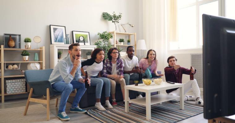 A diverse group of six friends excitedly watching TV together in a cozy living room, with a bowl of popcorn and drinks on the coffee table.