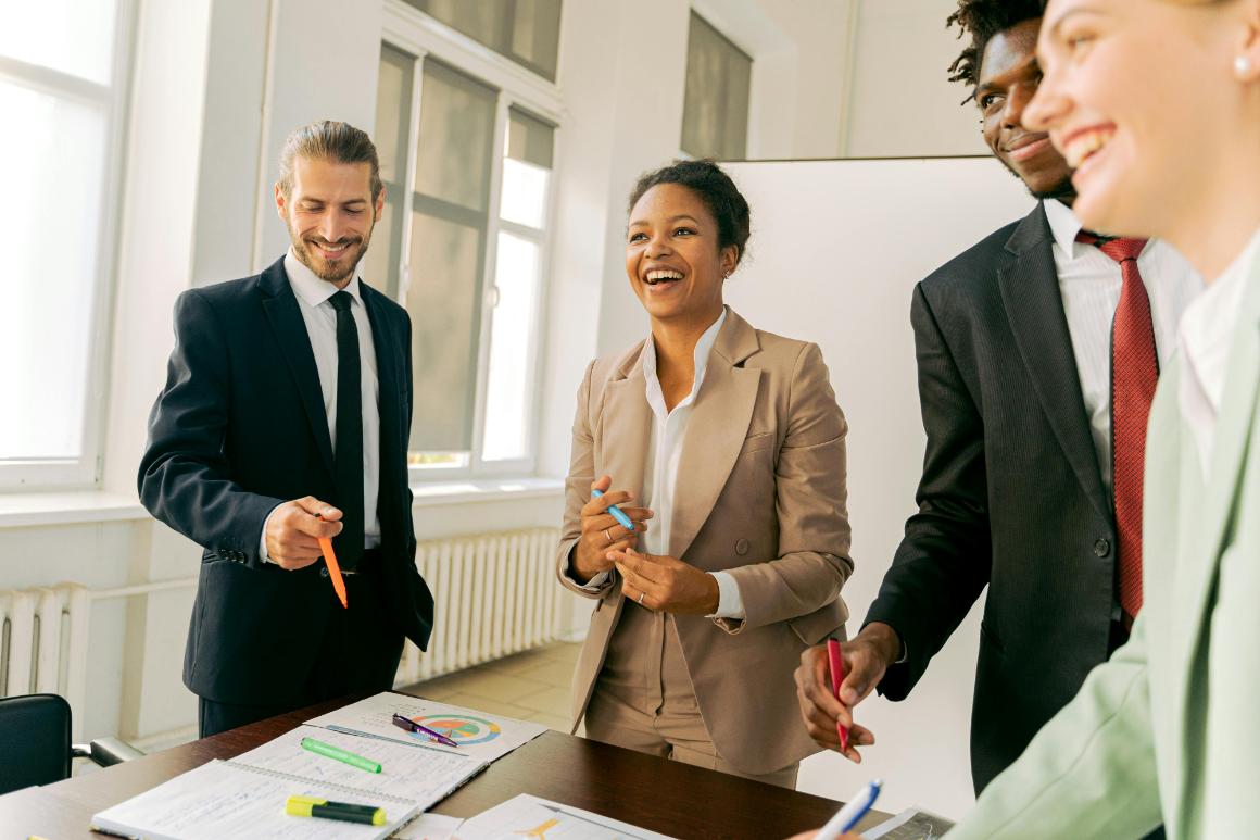 A group of four professionals, smiling and holding markers, engage in a collaborative discussion around a table with notebooks and charts in a bright office setting.