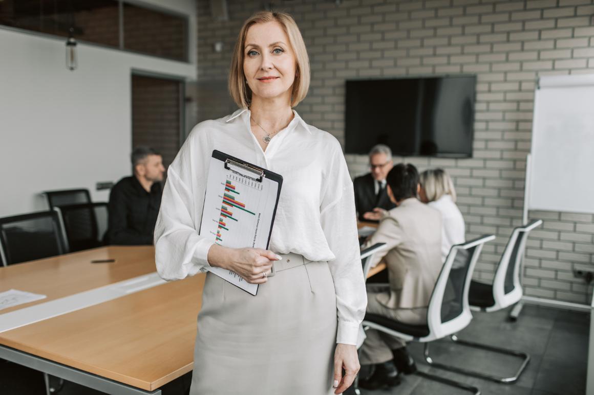 Woman holding a clipboard with a colorful bar chart in a modern office while coworkers meet in the background.