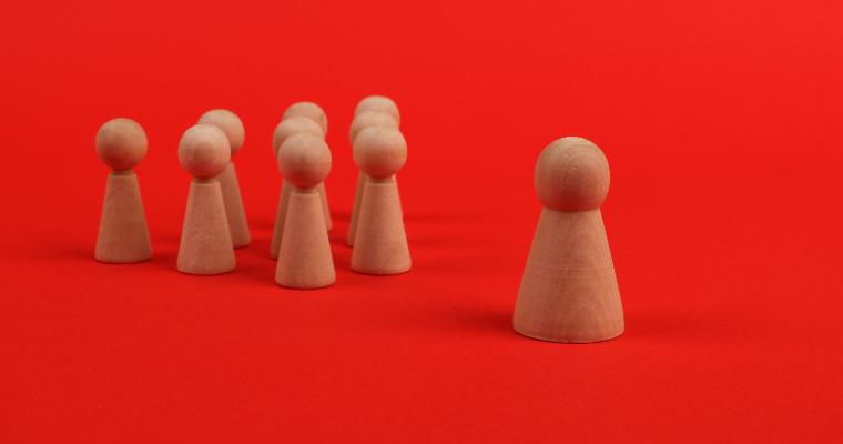 A group of wooden figurines on a red background, with one taller figure standing apart from six smaller figures. image link to article