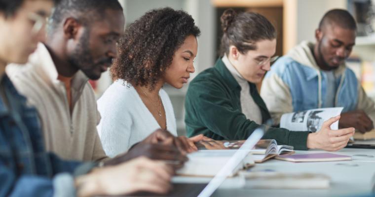A diverse group of five people seated at a table, focused on studying or reading materials in a library or study area.