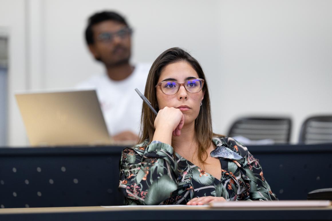 A woman in glasses attentively listens in a classroom, holding a pen and resting her chin on her hand