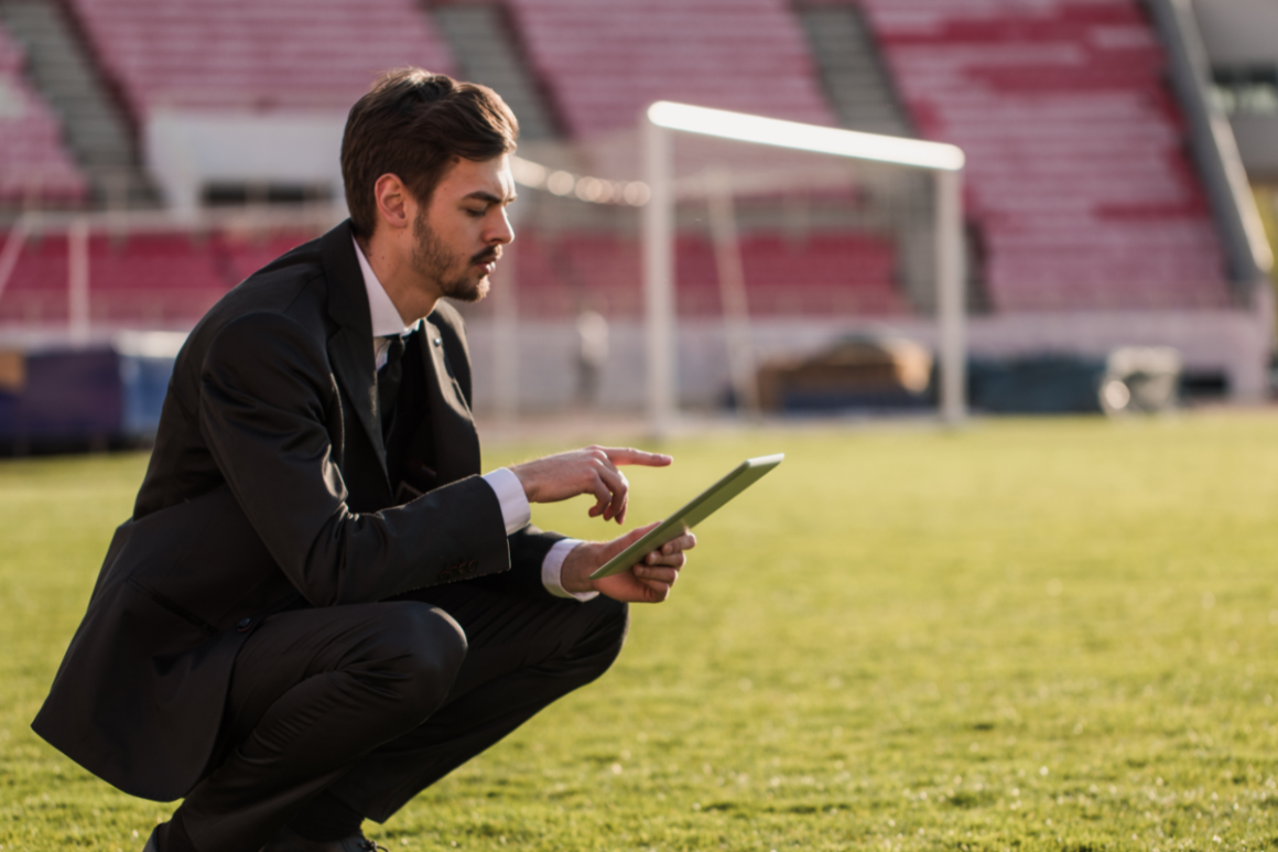 A man in a suit crouches on a soccer field, focused on a tablet
