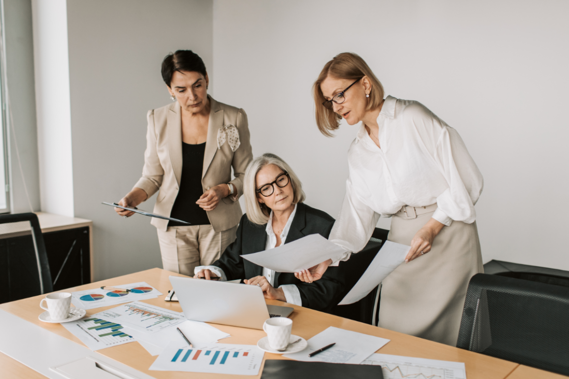 Three businesswomen review documents and charts around a conference table with an open laptop.
