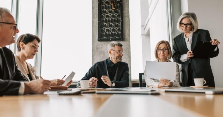 Five adults gather around a conference table with papers and folders in a bright office.