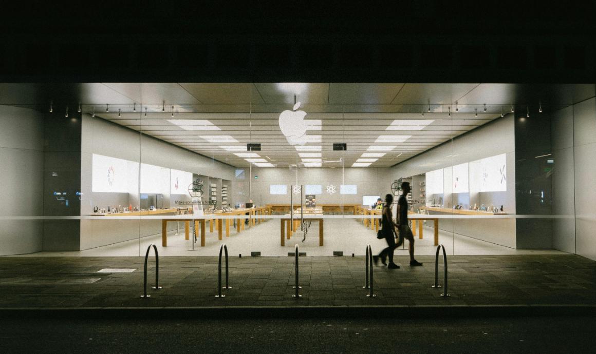 A brightly lit Apple store at night with large glass windows and wooden display tables. Two people walk past the store, and the Apple logo is visible above the entrance.