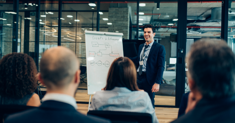 A presenter in a dark suit smiles while addressing an audience in a modern office. A whiteboard displays a flowchart titled Goals & Achievements.