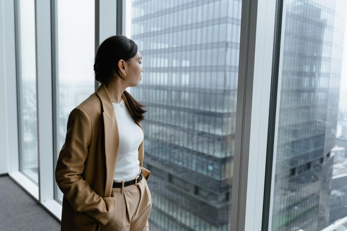 Woman in a tan suit standing by floor-to-ceiling windows and looking at surrounding city buildings.