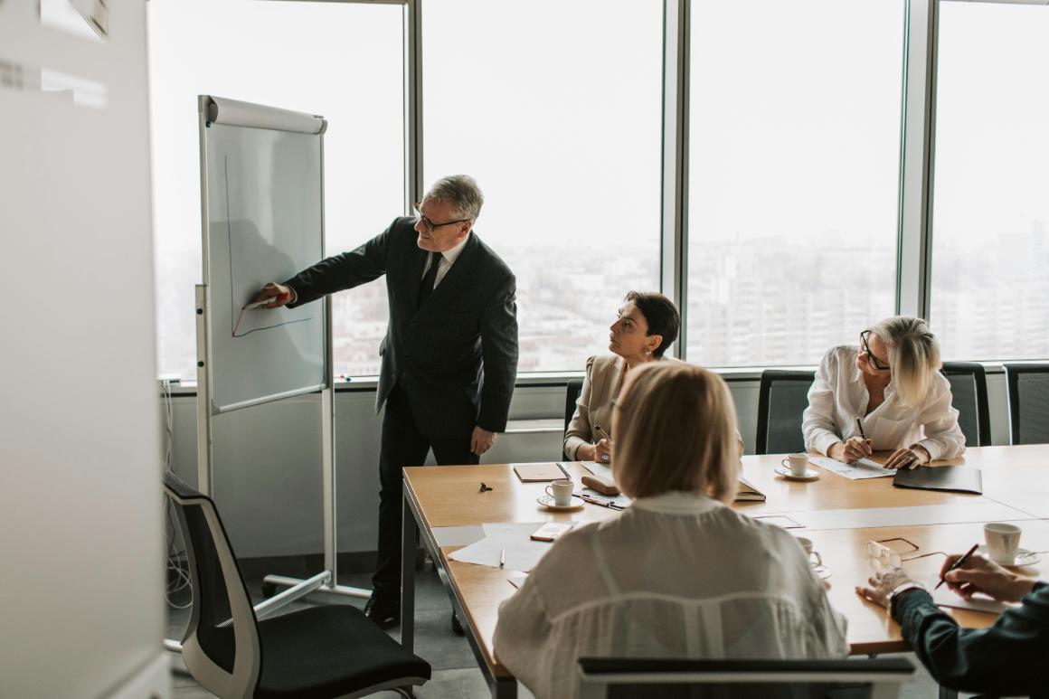 A man in a suit presents a graph on a flip chart to a group of three attentive women in a conference room with a city skyline view.