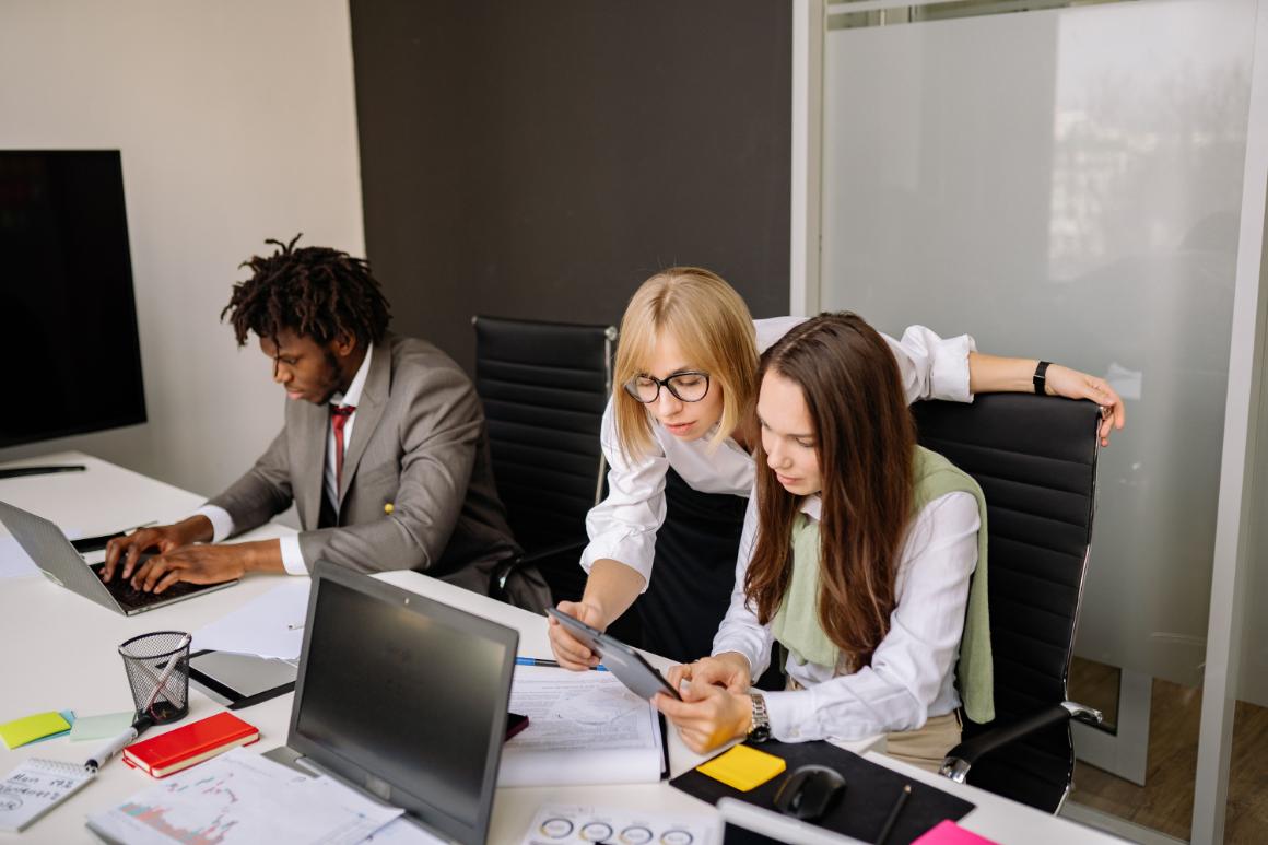 Three individuals collaborate in a modern office; a man types on a laptop while two women examine a tablet together.