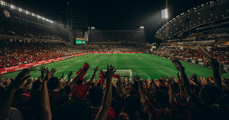 A vibrant football stadium at night, with fans in red jerseys cheering. The pitch is brightly lit, brimming with excitement and energy. image link to article