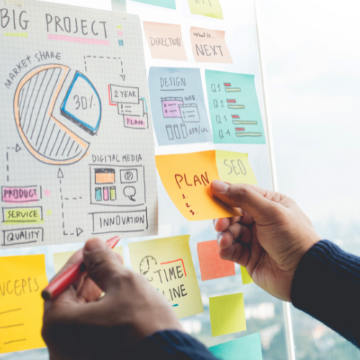 Close up of idea board with graphics, sticky notes, and hands reaching with a pen and new sticky note to add to the board. 