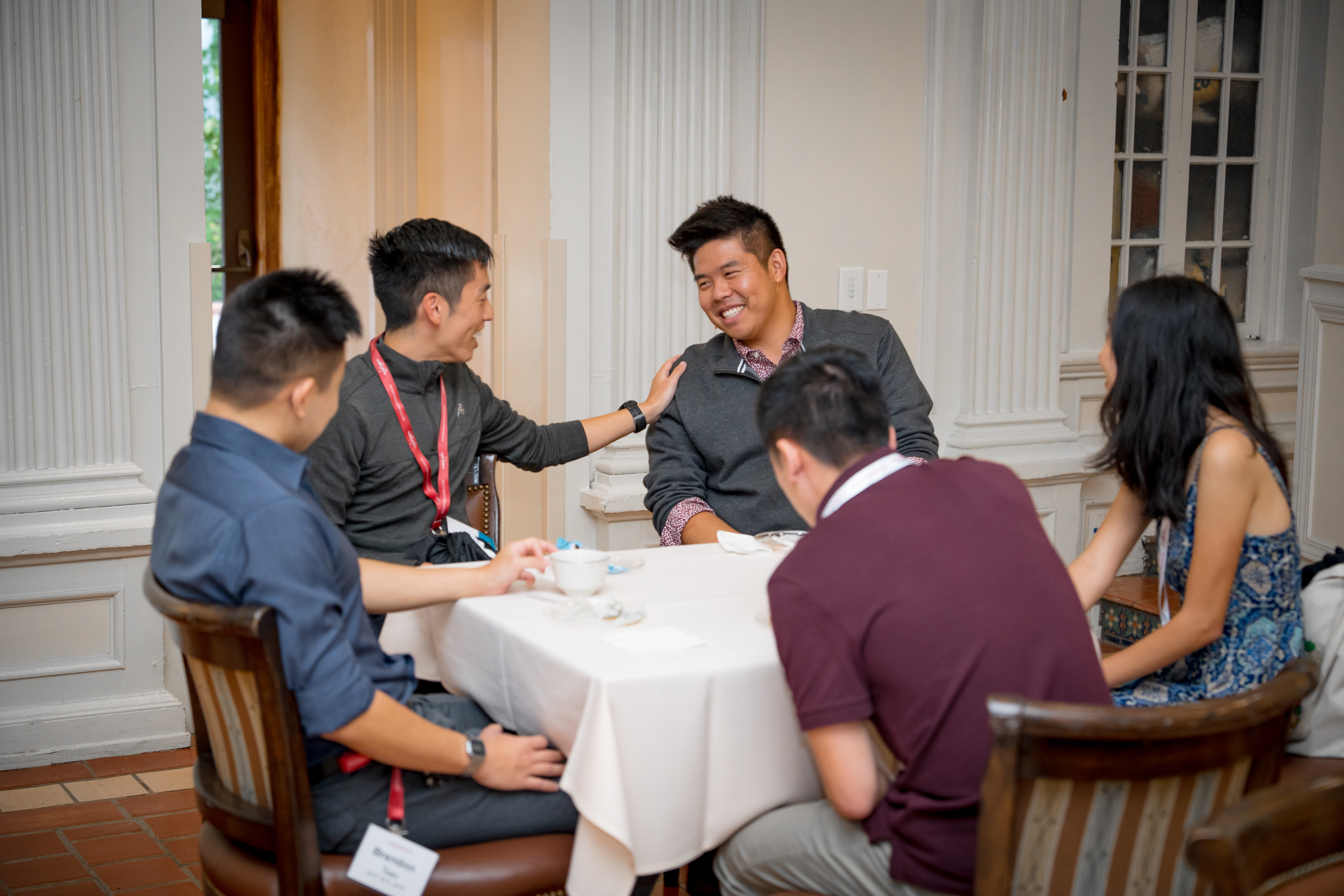 A group of five alumni seated around a table. One has his hand on the others shoulder, who is smiling.