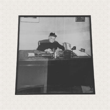 A framed black and white image of a Jesuit priest seated at a large wooden desk.