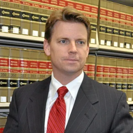 A man in a suit and tie looking off to the side while standing in front of a bookshelf full of law books.