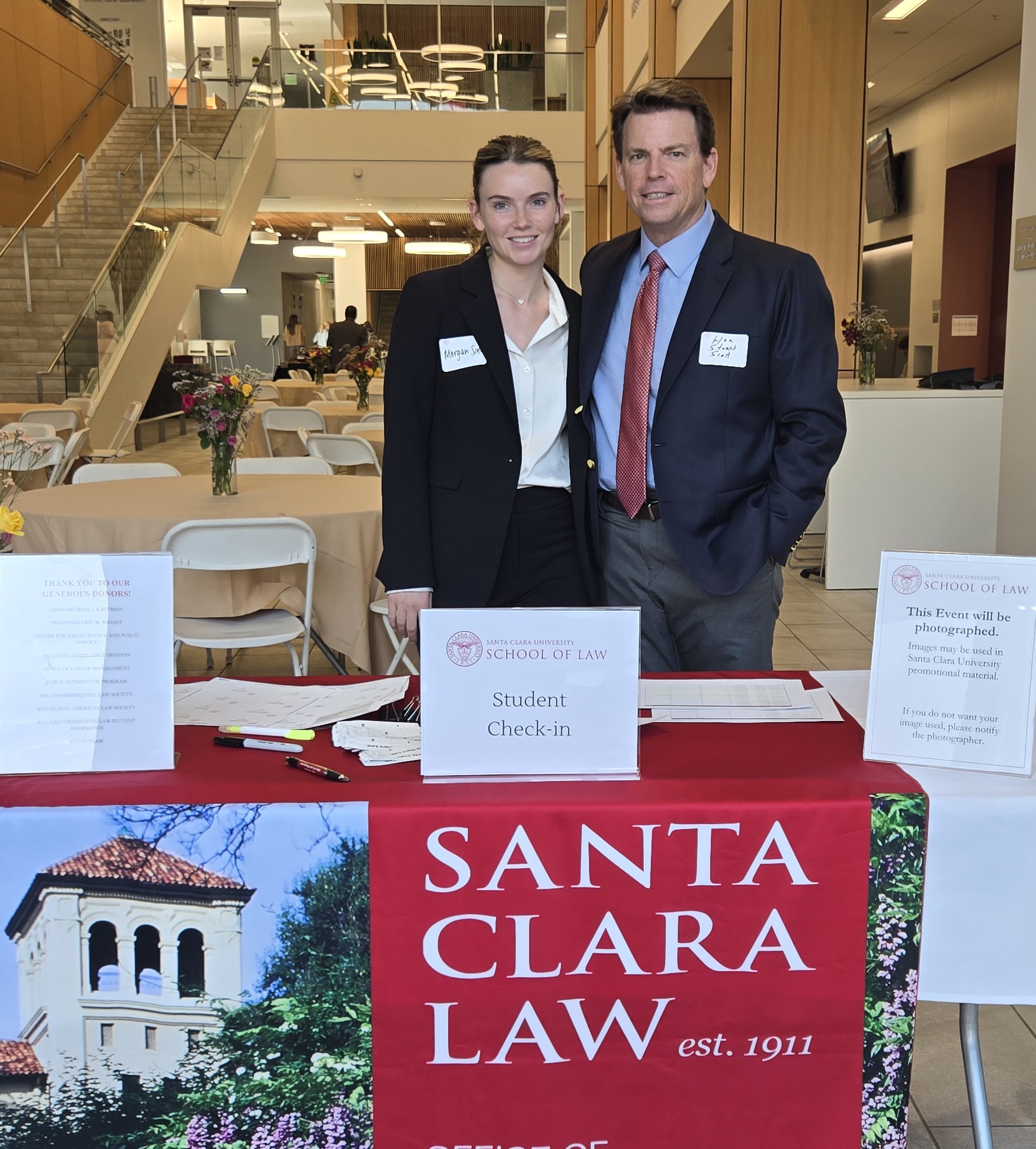 A man and a young woman in business wear standing behind an event check-in table with a banner reading Santa Clara Law. The mans nametag reads Hon. Stuart Scott and the young womans reads Morgan Scott.