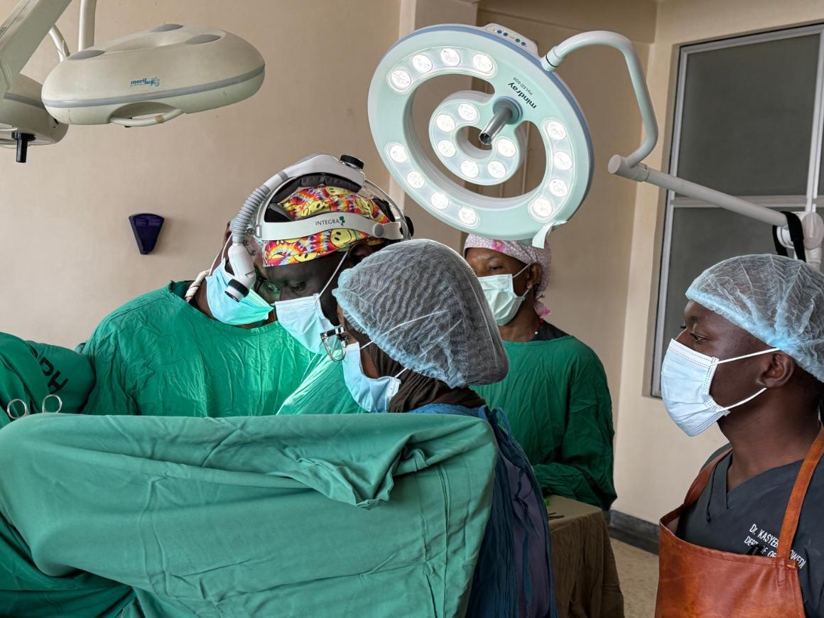 Several men and women in medical garb and hair nets stand under operating room lights. They appear to be mid-procedure, though the patient is not visible.