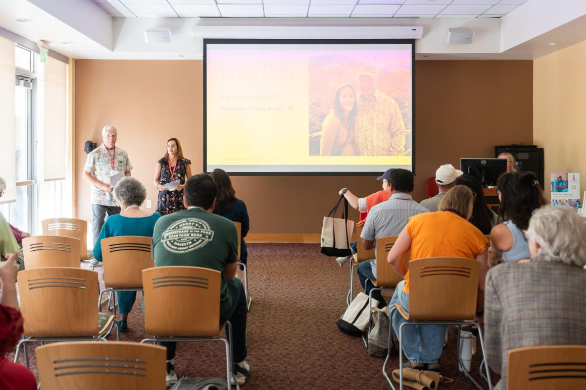 A man and a woman stand in front of a seated audience, and next to a projector screen with their photo on it, along with text reading Co-founders of The Musa Project. Hal Tilbury, Class of 65. Anastasia Tilbury Marks 95.