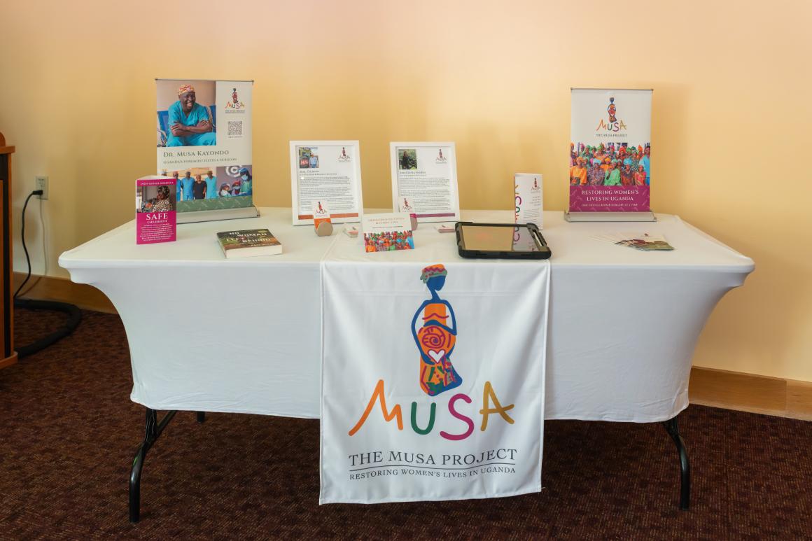 A table with a white tablecloth and a table runner reading Musa. The Musa Project. Restoring Womens Lives in Uganda. The tabletop holds several stacks of fliers and some small, pop-up banners with more information about The Musa Project.