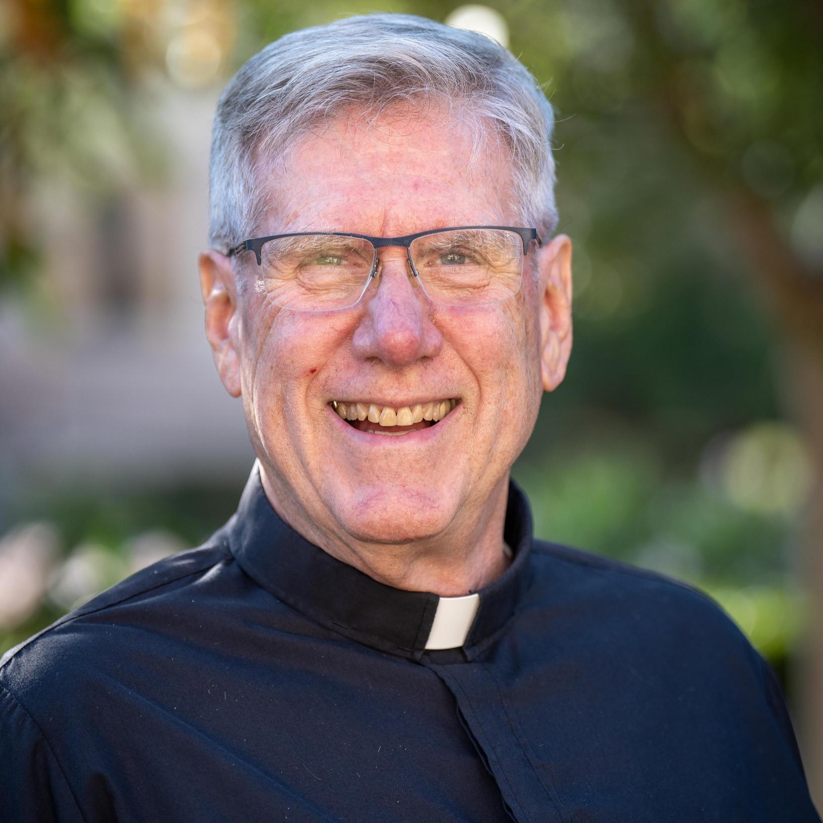 A man wearing a clerical shirt smiles in an outdoor setting. 