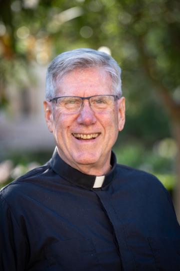 A man wearing a clerical shirt smiles in an outdoor setting. 