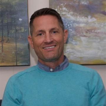 A person smiling in front of abstract paintings.