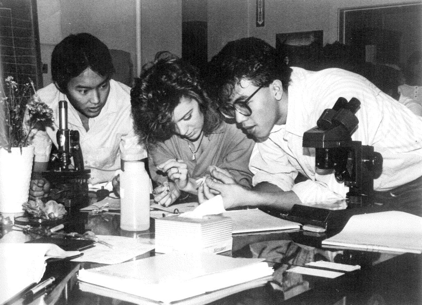 Three students examining documents in a lab with microscopes and equipment.