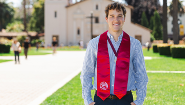 A young man in a red Santa Clara University graduation stole, standing in front of the Mission Church. 